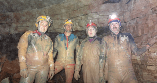 Four cavers in helmets and muddy suits pose inside a cave, surrounded by rocky walls.