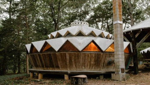 A unique, geometric wooden structure with a conical roof, surrounded by trees in a natural setting.