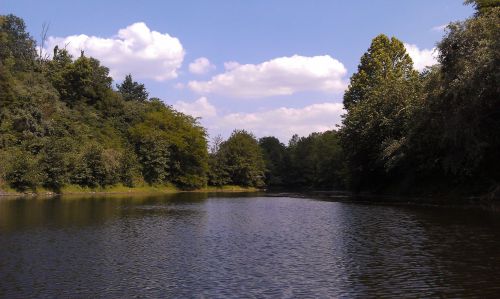 A serene river scene surrounded by lush greenery and trees under a blue sky with fluffy white clouds.