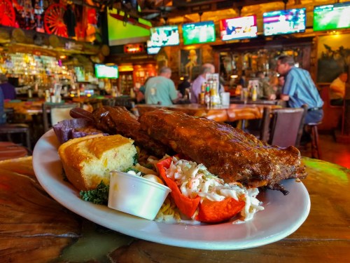 A plate of ribs with coleslaw and cornbread, set in a lively restaurant with people and TVs in the background.