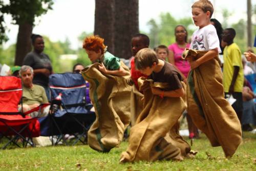 Three children in sack race bags run across a grassy field, with spectators in the background.