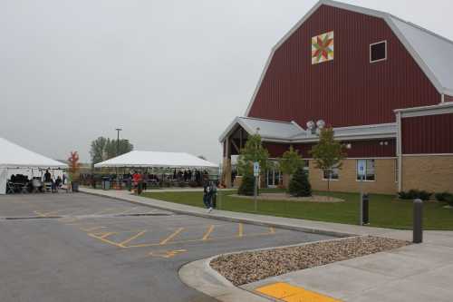 A red barn-style building with a quilt pattern, surrounded by tents and people on a cloudy day.