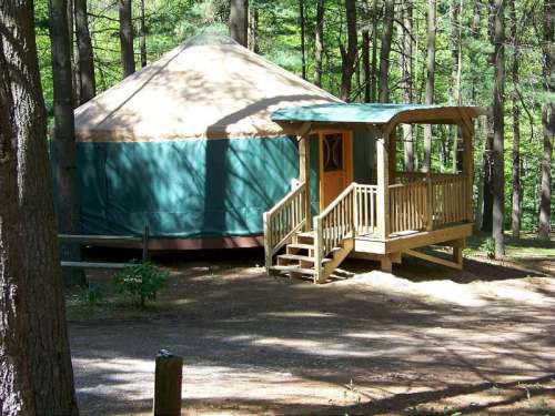 A yurt with a wooden porch nestled in a forested area, surrounded by trees and natural scenery.