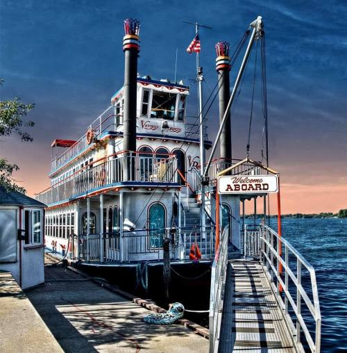 A colorful riverboat docked at a pier with a "Welcome Aboard" sign against a sunset sky.