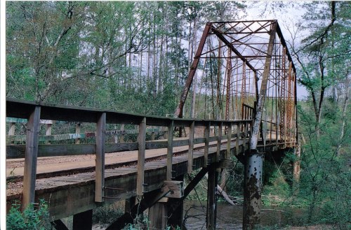 Rusty old bridge over a river, surrounded by lush green trees and a serene natural landscape.