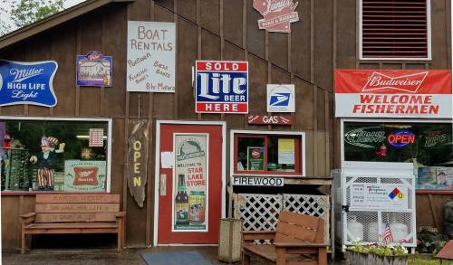 A rustic storefront with signs for boat rentals, beer, firewood, and welcoming fishermen, featuring a cozy bench outside.