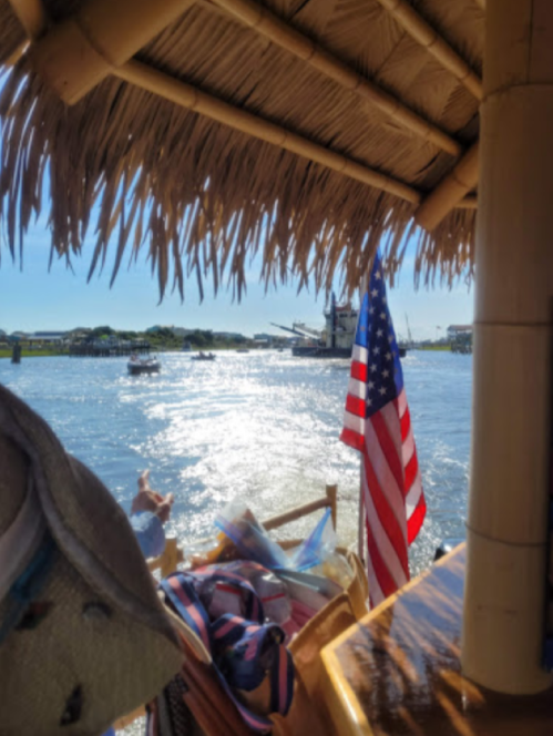 View from a tiki hut with an American flag, overlooking a sparkling waterway and boats in the distance.