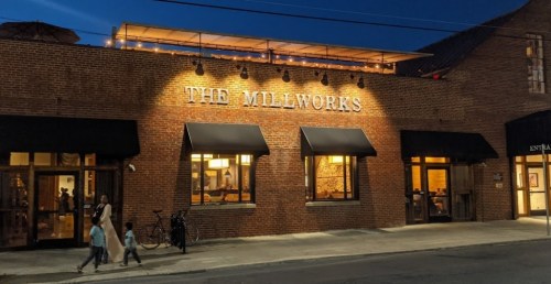 A brick building at night with warm lights, featuring "The Millworks" sign and people walking outside.