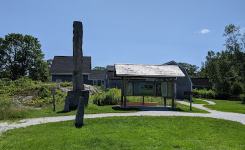 A grassy area featuring a tall totem pole, a wooden shelter, and a building in the background under a clear blue sky.