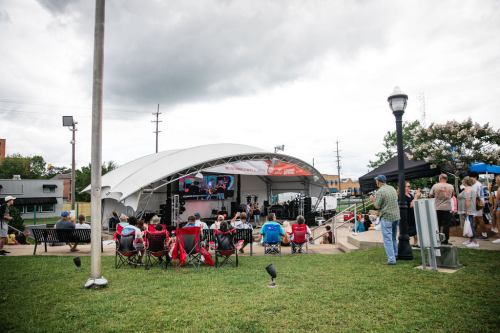 A crowd watches a performance on a stage under a cloudy sky, with people seated on lawn chairs in a park setting.