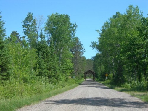 A gravel road lined with lush green trees leads to a covered bridge in a serene, natural setting.
