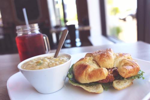 A croissant sandwich with lettuce and filling, served with a bowl of soup and a drink in a cozy café setting.