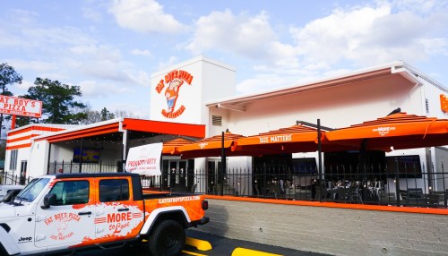 Exterior of a pizza restaurant with orange umbrellas, a white Jeep parked outside, and a clear blue sky.
