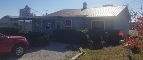 A small, blue restaurant with a red metal roof, surrounded by greenery and a sign that reads "Great Food."