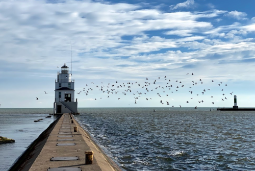 A lighthouse on a pier with a flock of birds flying over the water under a partly cloudy sky.