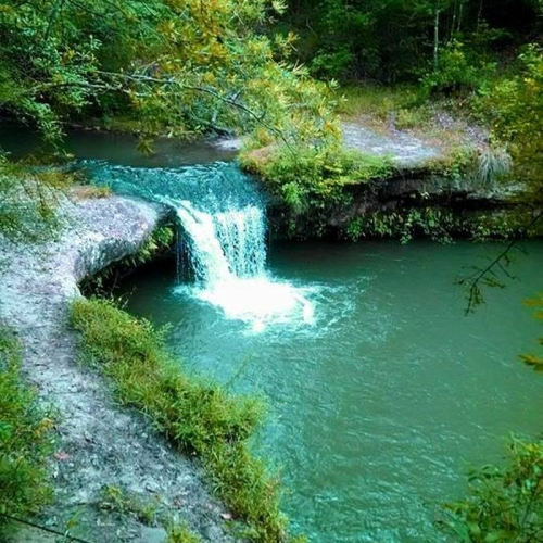A serene waterfall cascades into a tranquil green pool, surrounded by lush greenery and rocky terrain.