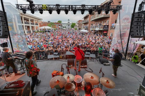A lively outdoor concert with a large crowd, featuring musicians on stage and colorful decorations.