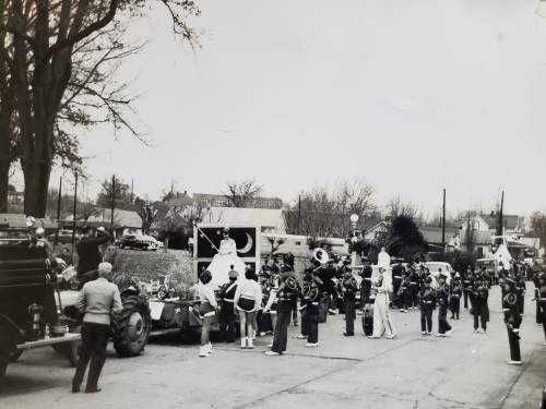 A black and white photo of a parade with a marching band, floats, and spectators along a street lined with trees.
