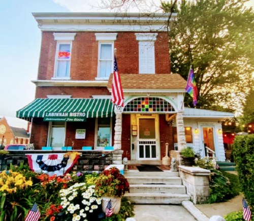 A charming bistro with a striped awning, flags, and colorful flowers in front of a brick building.