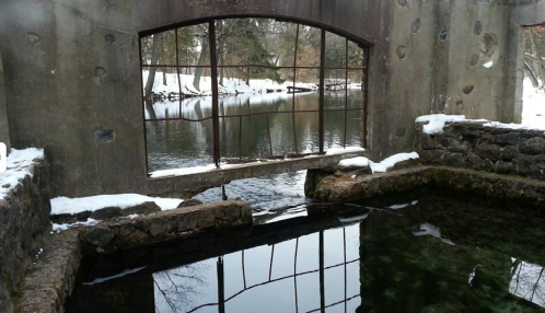 A snowy landscape with a stone structure and a calm, reflective pond under a large window frame.