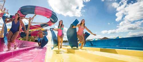 Four people enjoying a water park, carrying inflatable mats and running towards colorful water slides under a sunny sky.