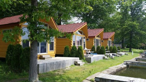 Four yellow cabins with red roofs line a grassy area by a calm lake, surrounded by trees and shrubs.