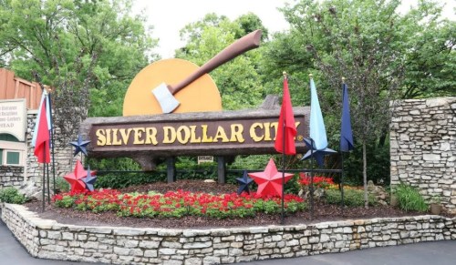 Sign for Silver Dollar City with an axe, surrounded by colorful flags and flowers, set against a green backdrop.