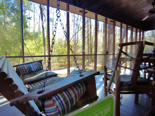 A screened porch with wooden rocking chairs and cushions, surrounded by trees and natural light.