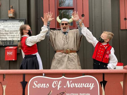 Three people in Viking costumes celebrate with raised hands in front of a "Sons of Norway" sign. All are wearing masks.