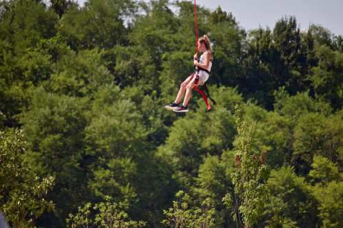 A person in a harness swings from a rope above a lush green landscape on a sunny day.