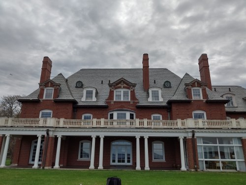 A large brick mansion with a gray roof, featuring multiple chimneys and a spacious porch, set against a cloudy sky.