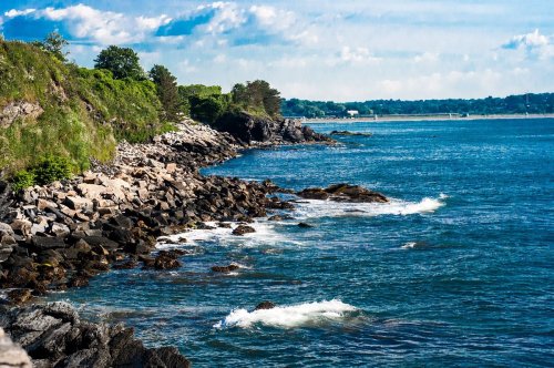 Rocky shoreline with waves crashing, lush greenery in the background, and a clear blue sky above.
