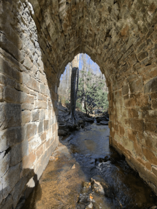 A stone archway over a stream, surrounded by trees and greenery, with sunlight filtering through.