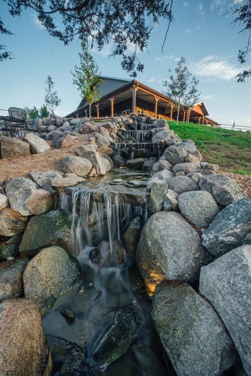 A serene waterfall flows over rocks, leading to a modern building surrounded by trees and a clear blue sky.