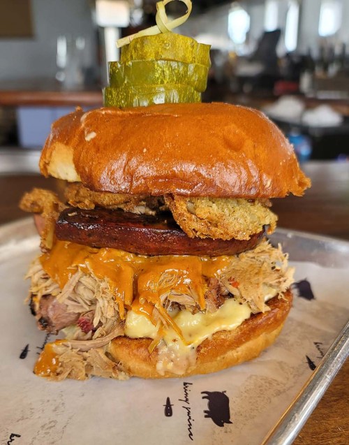 A stacked burger with pulled pork, cheese, fried pickles, and a pickle on a toasted bun, served on a tray.