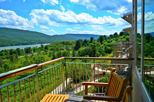 A scenic view from a balcony overlooking a river, lush greenery, and mountains under a bright blue sky with clouds.