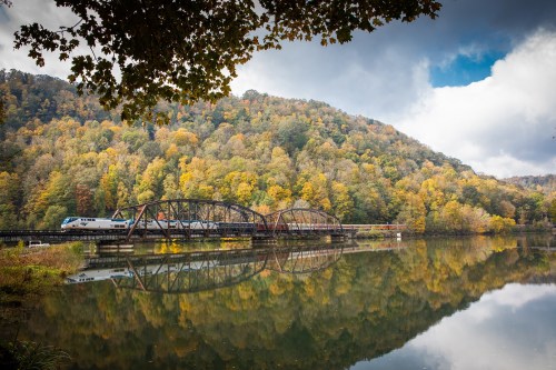 A train crosses a bridge over a calm river, surrounded by autumn-colored trees and a cloudy sky.