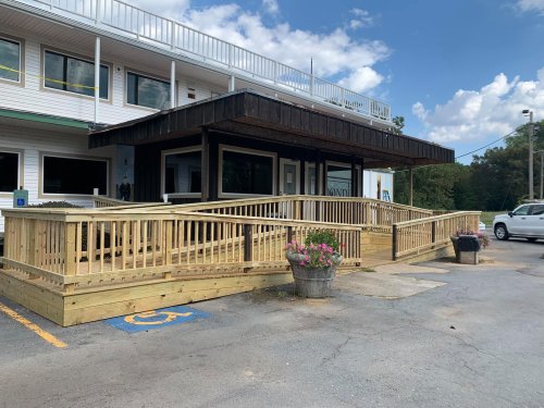 A wooden ramp leads to a building entrance, with flower pots and a parking area visible under a blue sky.