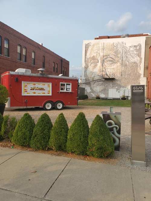 A red food truck parked near a large mural of a face on a brick building, with neatly trimmed hedges in the foreground.