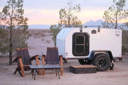 A compact white trailer parked in a desert setting, with two wooden chairs and a table nearby, surrounded by trees.