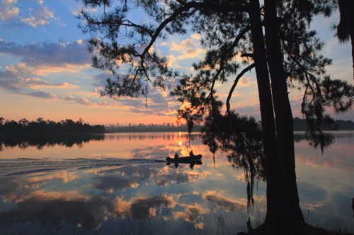 A serene lake at sunset, with a silhouette of a boat and trees reflecting on the calm water.