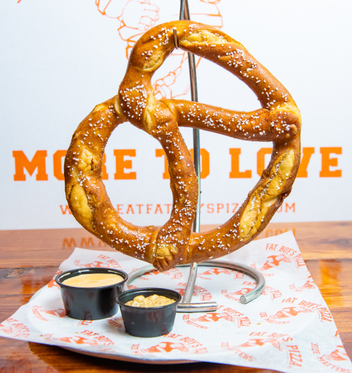 A large, soft pretzel on a stand, sprinkled with salt, accompanied by two dipping sauces on a branded paper.