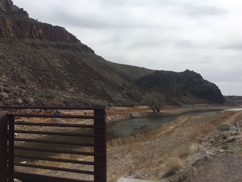 A serene landscape featuring a river bordered by rocky hills and sparse vegetation under a cloudy sky.