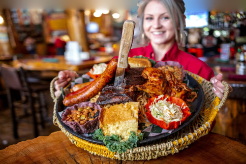 A smiling server holds a large platter of assorted barbecue meats, sides, and cornbread in a rustic restaurant setting.
