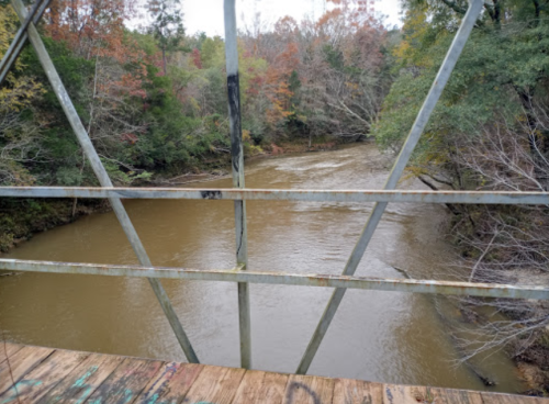 View of a muddy river flowing beneath a metal bridge, surrounded by trees with autumn foliage.