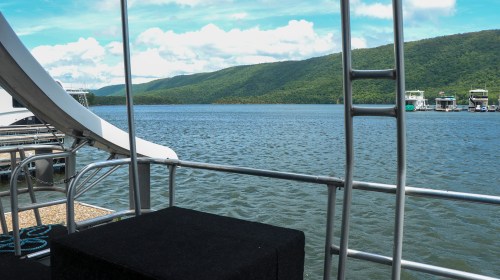 View of a calm lake surrounded by green hills, with boats docked in the distance under a blue sky.