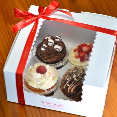 A box of four decorated cupcakes, featuring chocolate, strawberry, and raspberry toppings, tied with a red ribbon.
