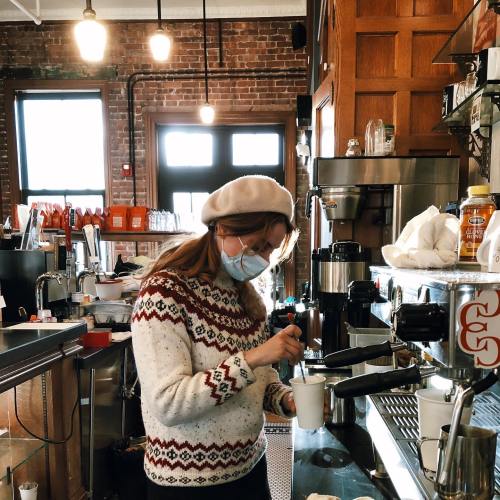 A barista in a cozy café prepares a drink, wearing a mask and a festive sweater, with coffee equipment in the background.