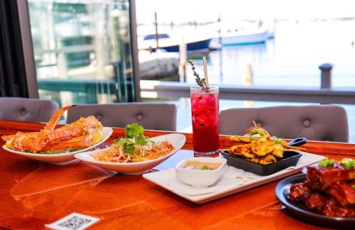 A table with seafood dishes, a cocktail, and a waterfront view in the background.
