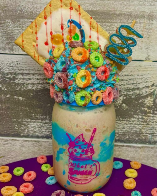 A colorful milkshake topped with cereal, a pastry, and candy, served in a jar with a playful design.